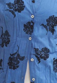 Chemise bleu à boutons avec motifs floraux noirs, texture lisse et boutons décoratifs. Points forts incluent des dessins contrastants de pétales et de feuilles.
