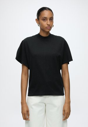 Vrouw met strak naar achter gekamd haar draagt een los zwart T-shirt, witte broek en gouden hoepeloorbellen, staand tegen een effen lichte achtergrond.