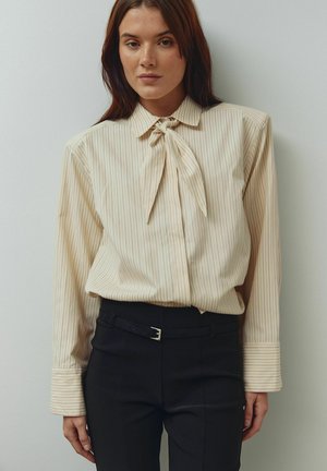 Beige raidallinen pusero, jossa kauluksessa rusetti, pitkät hihat ja leveät kalvosimet, yhdistettynä mittatilaustyönä tehtyihin mustiin housuihin, joissa hopeinen solki.