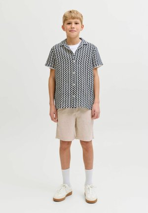 Garçon debout portant une chemise à manches courtes à carreaux noir et blanc, un short beige, des chaussettes blanches et des baskets blanches sur un fond uni.