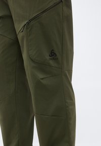 Közeli kép olívazöld színű kültéri nadrágról cipzáras zsebbel és fekete Odlo logóval a combon.