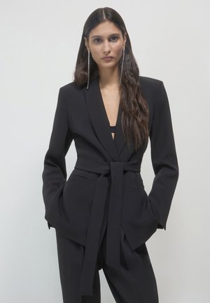 Blazer noir ajusté avec un décolleté en V plongeant et une ceinture à nouer, doté de poches latérales et d'épaules structurées. Le modèle porte de longues boucles d'oreilles scintillantes.