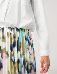 Faltenrock aus mehrfarbigem Stoff mit abstrakten Mustern in Grün, Blau und Pink; kombiniert mit einem lockeren weißen Hemd mit Knöpfen.