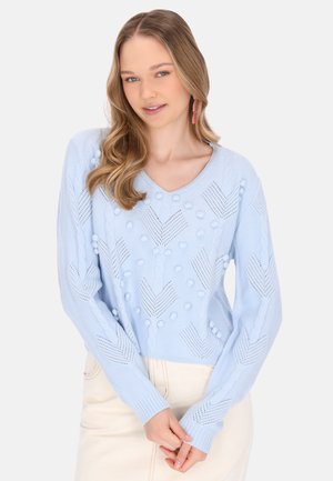 Femeie cu păr lung și blond poartă un pulover tricotat albastru deschis, texturat și o fustă crem, stând cu mâinile încrucișate în față.