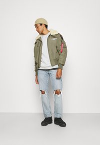 Olivgrüne Bomberjacke mit Kunstfellkragen, bestickten Patches und roten Stoffakzenten, kombiniert mit hellblauen, zerrissenen Jeans und schwarzen Schuhen.