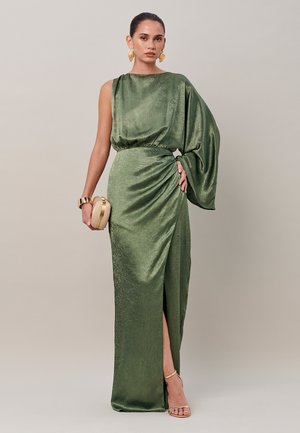 Femme portant une robe longue au sol, brillante, vert olive à une épaule avec une manche drapée, tenant une pochette beige et portant des boucles d’oreilles et un bracelet dorés.