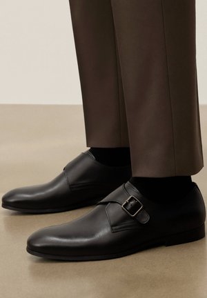 Schwarze Ledermokassins mit Riemen, getragen zu dunkelbraunen Hosen und schwarzen Socken, stehend auf beigem Boden.