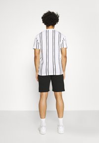 Bílé pruhované tričko s černými proužky, spárované s černými šortkami. Tričko má logo jako ozdobu, umístěné na jednotném bílém pozadí.