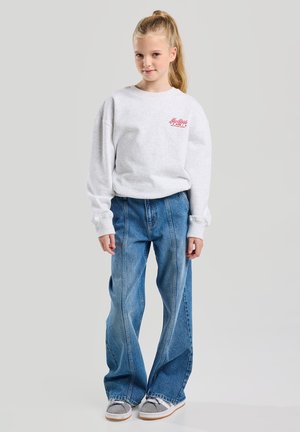 Jeune fille avec une queue de cheval blonde portant un sweat-shirt gris clair, un jean bleu large et des baskets grises, debout devant un fond uni.