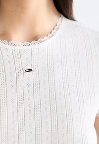 Biały sweter z dzianiny z pionowymi paskami, falbaniastym dekoltem z koronki oraz małym logo marki na środku. Tekstura z delikatnymi wzorami.