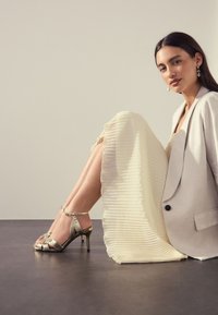 Sandali con tacco alto color argento metallico con cinturini incrociati e fibbia alla caviglia, abbinati a un vestito plissettato avorio e a un blazer grigio chiaro.