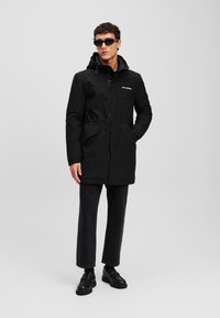Sort parka-jakke med lynlås foran, snoretræk i livet og hætte. Har sidelommer og logo på brystet. Bæres med sorte jeans og støvler.