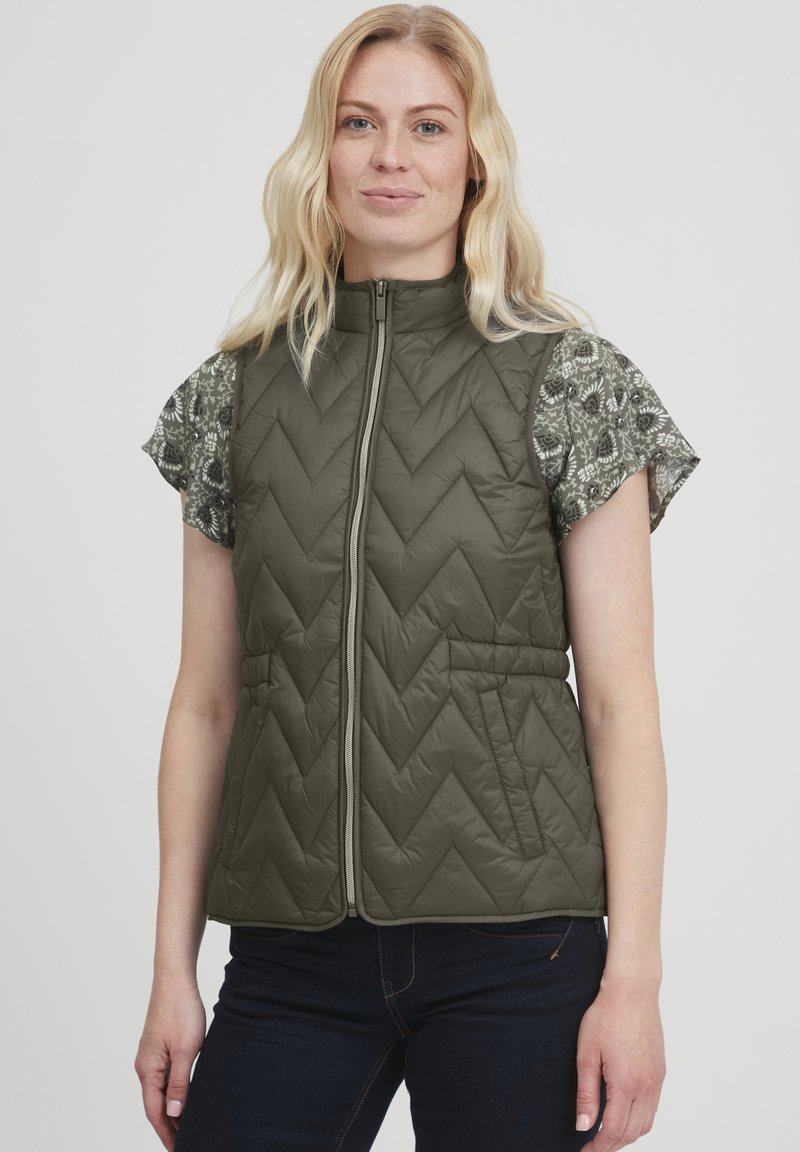 Chaleco acolchado de color verde oliva con un patrón de espiga, cuello alto, cremallera frontal y bolsillos laterales. Lleva puesto un top de manga corta con estampado.