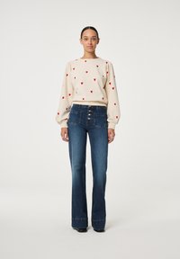 Hellbeige Pullover mit roten Herzmustern, kombiniert mit dunkelblauen Schlagjeans mit hoher Taille und Knopfverschluss. Sanfte Texturen.