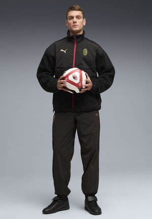 Jonge man in een zwart Puma trainingspak houdt een rood-witte AC Milan voetbal vast tegen een grijze verloopachtergrond.