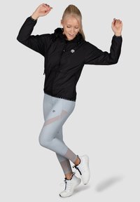 Sort letvægtsjakke med hætte, der har elastiske manchetter og snor. Lys grå leggings med udskårne detaljer, parret med hvide sneakers.