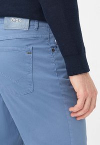 Hellblaue Hosen mit glatter Textur, ausgestattet mit einer Gesäßtasche mit Brax-Label, geradem Schnitt und kontrastierenden Nähdetails.