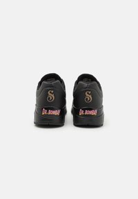Schwarze Sneaker mit glattem Obermaterial, das ein Logo und den Text "DR. BOMBAY" in einem rosa Farbverlauf an der Ferse präsentiert. Gummisohle mit strukturiertem Grip.