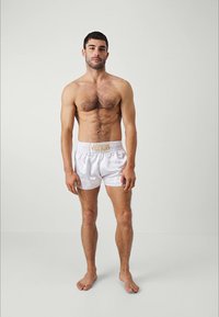 Muscular man stands barefoot wearing white Venum branded shorts against plain white background, arms relaxed by his sides.