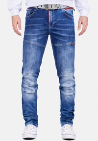 Jean en denim dans un délavage bleu moyen avec des sections usées, présentant des détails de couture, des accents en patch et une poche zippée pour une fonctionnalité supplémentaire.