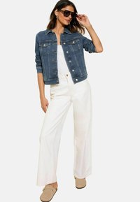 Giacca di jeans blu scuro con chiusura a bottoni, due tasche sul petto e maniche arrotolate. Abbinata a pantaloni bianchi a gamba larga e slip-on beige.