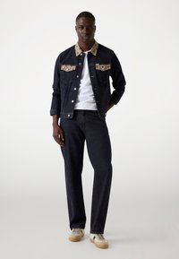 Chaqueta de mezclilla oscura con cuello y bolsillos estampados, combinada con pantalones de mezclilla oscura y zapatillas beige con acentos verdes.