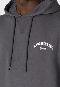 Felpa grigia con cappuccio e tasca a marsupio frontale, con coulisse e scritta bianca "SPORTING Goods" sul petto. Tessuto morbido in misto cotone.