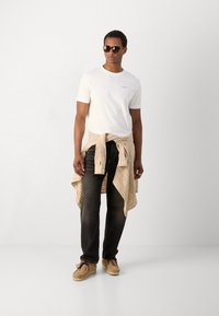 Witte T-shirt met korte mouwen, beige geblokte overhemd om de taille geknoopt, donkere jeans en lichtbruine schoenen. Model draagt een zonnebril.