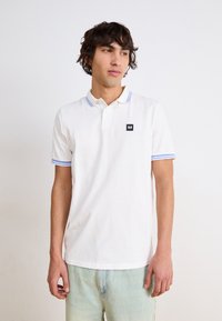 Hvid polo skjorte med blåstribede kraver og manchetter, har et lille sort logo patch på venstre bryst, lavet af bomuldsstof.