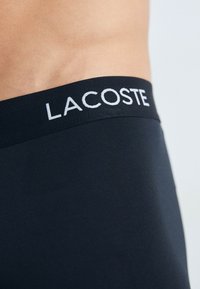 Boxers en coton noir avec une ceinture élastique texturée portant le logo « LACOSTE » en blanc. Tissu lisse avec un ajustement proche.