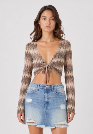 Kobieta ubrana w brązowy crop top z długim rękawem i wzorem w zygzak oraz zawiązywaną z przodu oraz jasno-niebieską, przystrzępioną mini spódnicę z denimu.