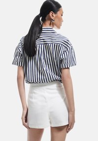 Camisa de manga corta con rayas verticales en azul marino y blanco, combinada con pantalones cortos blancos de cintura alta. Presenta un corte relajado y detalle de yugo en la parte trasera.