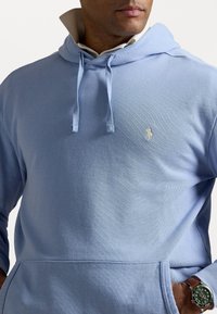 Man wearing light blue hoodie with drawstrings, white collared shirt underneath, and green wristwatch on left wrist, hand in pocket.