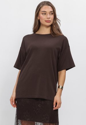 Vrouw draagt een losse donkerbruine top met korte mouwen en een zwarte kanten rok, met een gouden armband en kleine creolen, kijkt opzij.