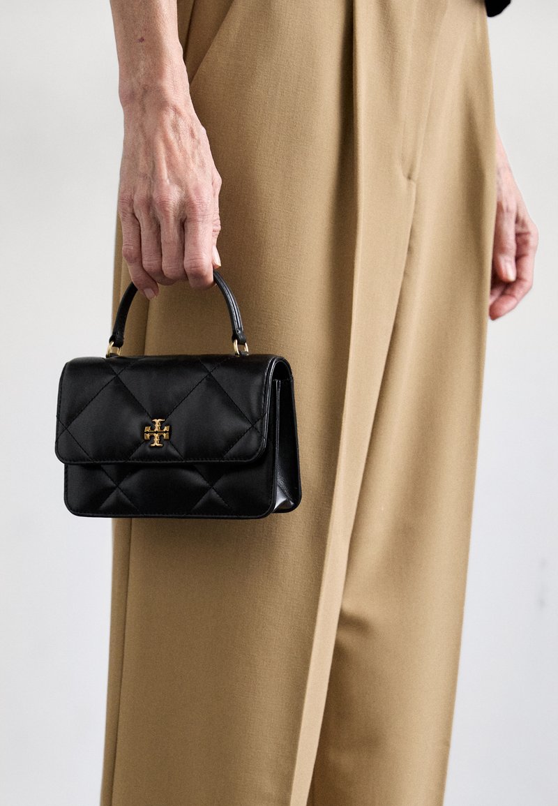 Svart läderhandväska med quiltat mönster, som har ett handtag högst upp och guld-tonade detaljer, hålls av en person i beige byxor.