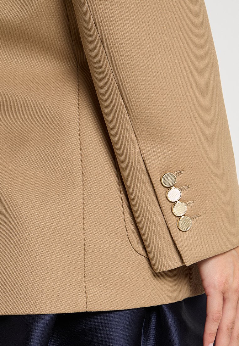 Beige op maat gemaakte blazer met een gestructureerd ontwerp, voorzien van een soepele textuur, zichtbare stiksels en drie gouden knopen als accent op de manchet.