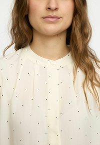 Blusa color crema con pois neri, con colletto alto, parte frontale plissettata e chiusura con bottoni. Tessuto morbido e leggero.