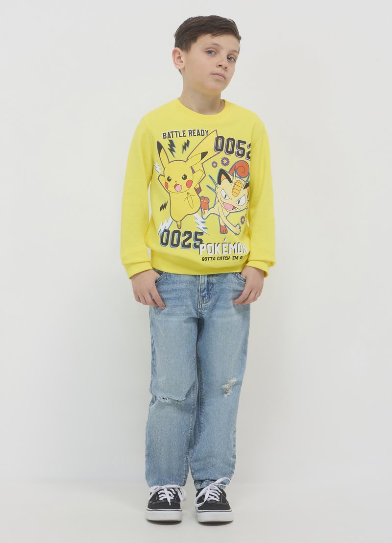 Gelbes langärmliges Sweatshirt mit Pikachu- und Meowth-Grafiken, kombiniert mit hellblauen, zerfetzten Jeans und schwarzen Sneakers mit weißen Schnürsenkeln.