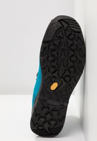 Türkisfarbene Wildlederschuhe mit einer schwarzen Gummisohle, die ein strukturiertes Grip-Muster und ein braunes Vibram-Logodetail an der Unterseite aufweisen.