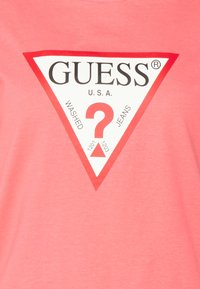 Korale T-shirt z dużą białą grafiką w kształcie trójkąta, czarnym logo "GUESS", czerwonymi akcentami oraz centralnym symbolem '?' z napisem "WASHED JEANS."
