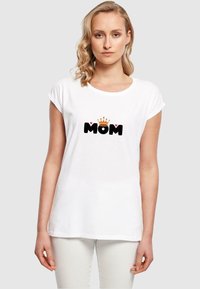 Weißes T-Shirt mit umgeschlagenen Ärmeln, das den schwarzen Text "MOM", eine goldene Krone darüber und kleine pinke Herzen auf beiden Seiten zeigt. Aus Baumwollmaterial.