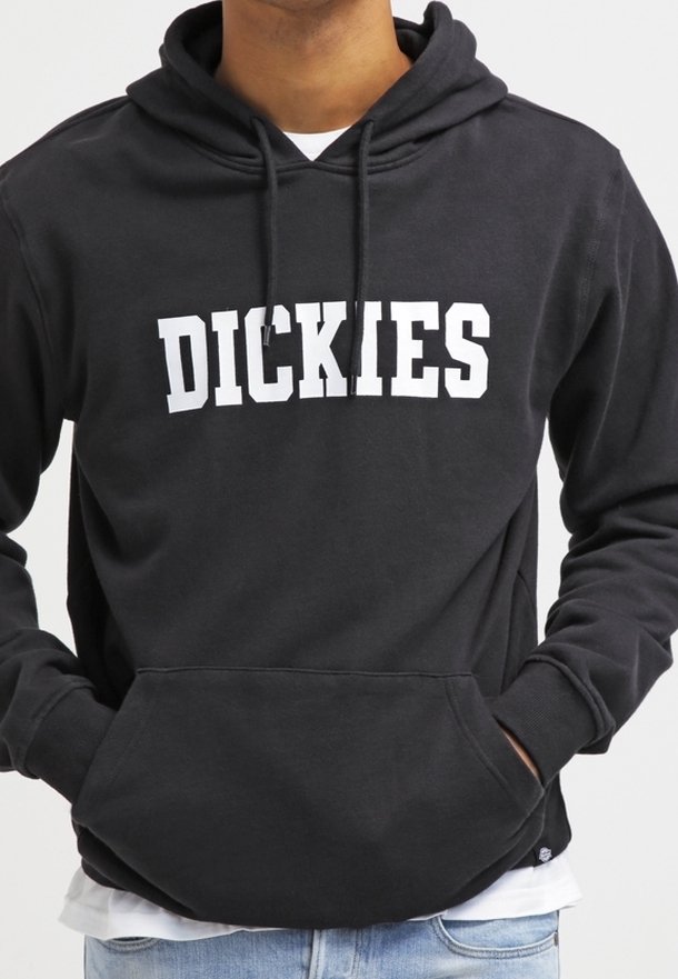 Černá mikina s kapucí a bílým logem "DICKIES" na hrudi, kterou nosí osoba se zvednutými rukama v přední kapse, v kombinaci s světle modrými džíny a bílým tričkem.