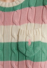 Gros plan d'un pull en tricot avec de larges rayures horizontales vertes, roses et crème, comportant une petite poche avant avec un logo brodé rose.