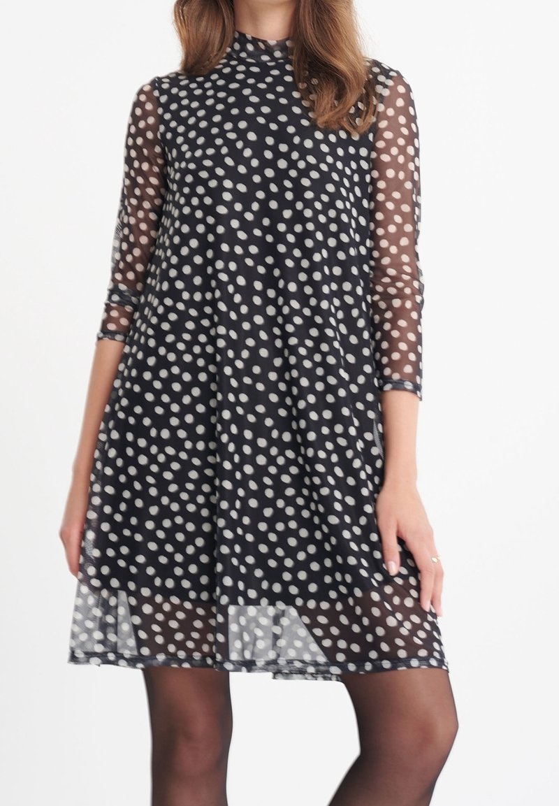 Femme portant une robe noire mi-longue à pois blancs avec des manches trois-quarts transparentes, assortie à des collants noirs.