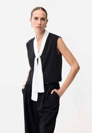 Femme aux cheveux lissés en arrière, portant un haut noir sans manches avec une cravate blanche autour du cou et un pantalon noir, les mains dans les poches, expression neutre.