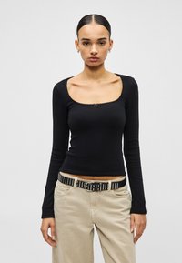 Juoda ilgomis rankovėmis palaidinė su kvadratiniu iškirpimu, centre puošta maža kaspino detale. Derinama su šviesiai smėlio spalvos kelnėmis ir juodu diržu su kaiščiais.