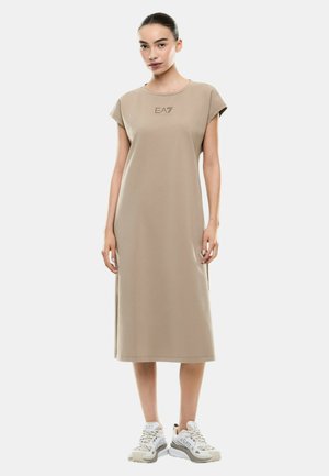 Donna in piedi che indossa un vestito midi beige EA7 a maniche corte abbinato a sneakers bianche EA7 su sfondo uniforme.