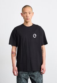 Camiseta de algodón negra con cuello redondo, mangas cortas y un logo circular en el pecho. Presenta un corte holgado y una textura suave.