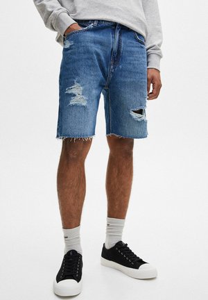 Homme portant un short en denim bleu déchiré, un sweat-shirt gris, des chaussettes blanches et des baskets noires, debout avec une main dans la poche.