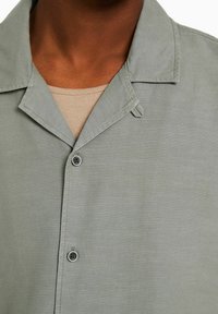 Jasnoszara koszula zapinana na guziki z kołnierzykiem typu spread, teksturowana tkanina i wyraźny detal guzika. Nałożona na beżowy, prążkowany top.